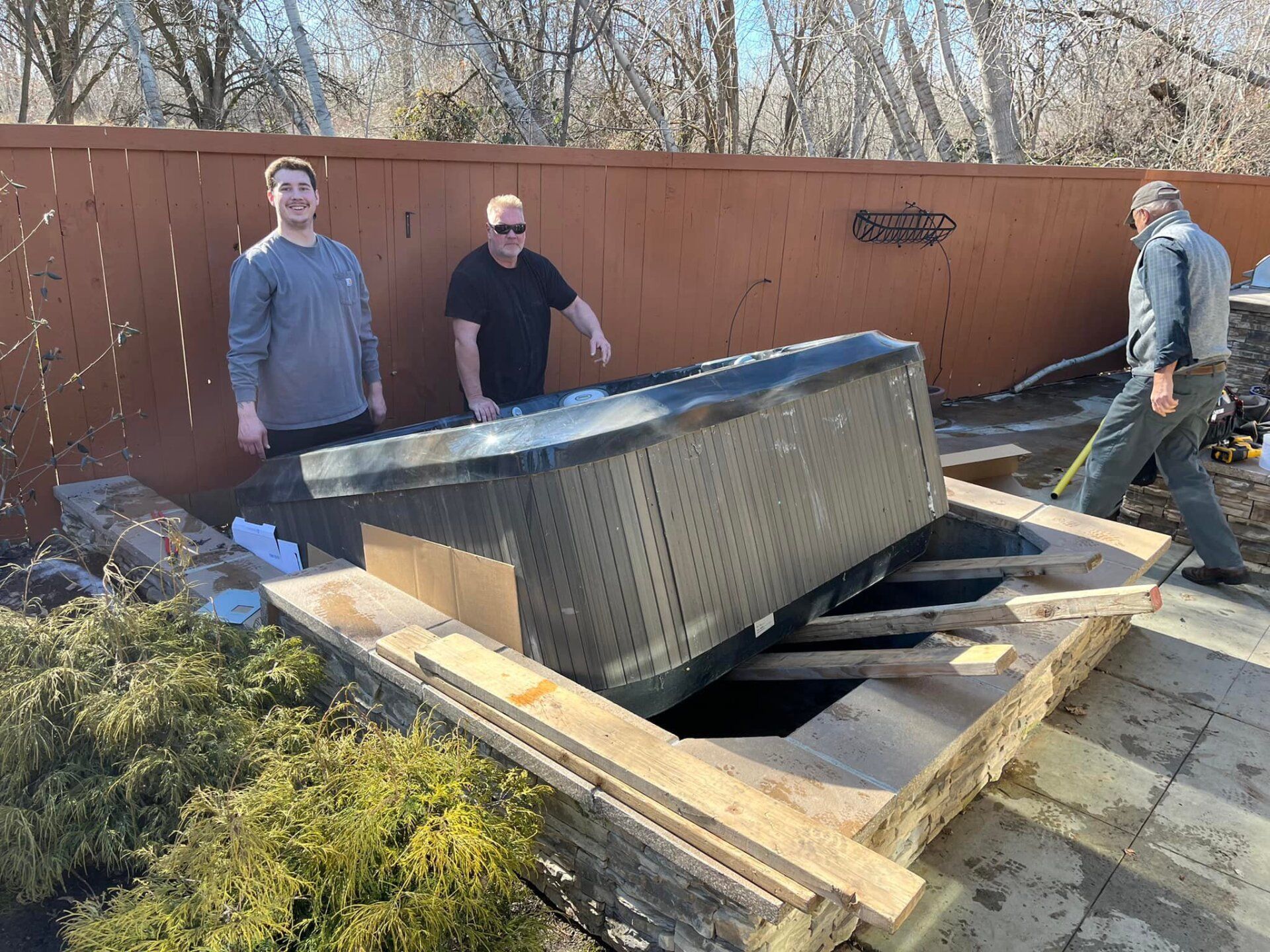 Three people dismantling a hot tub on a patio. Wooden frame and fence in background.