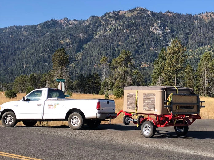 White pickup truck towing a trailer with a brown box, mountain backdrop.