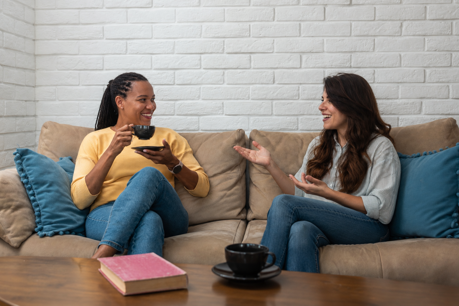 Two people chatting on a couch in a cozy living room, holding cups and smiling.