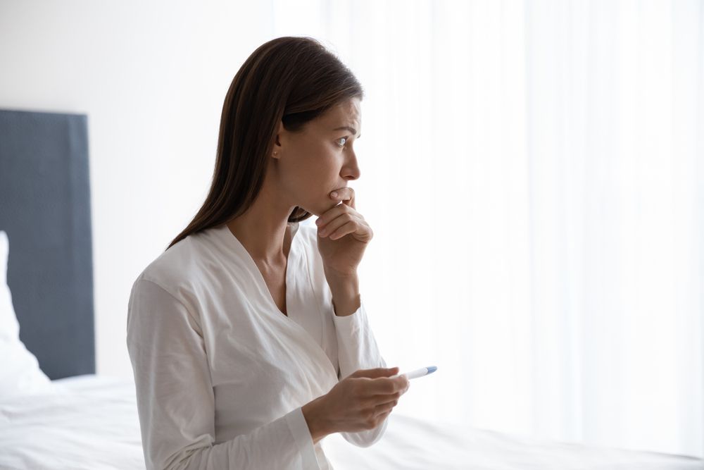 A concerned young woman looking out her window while holding a pregnancy test.