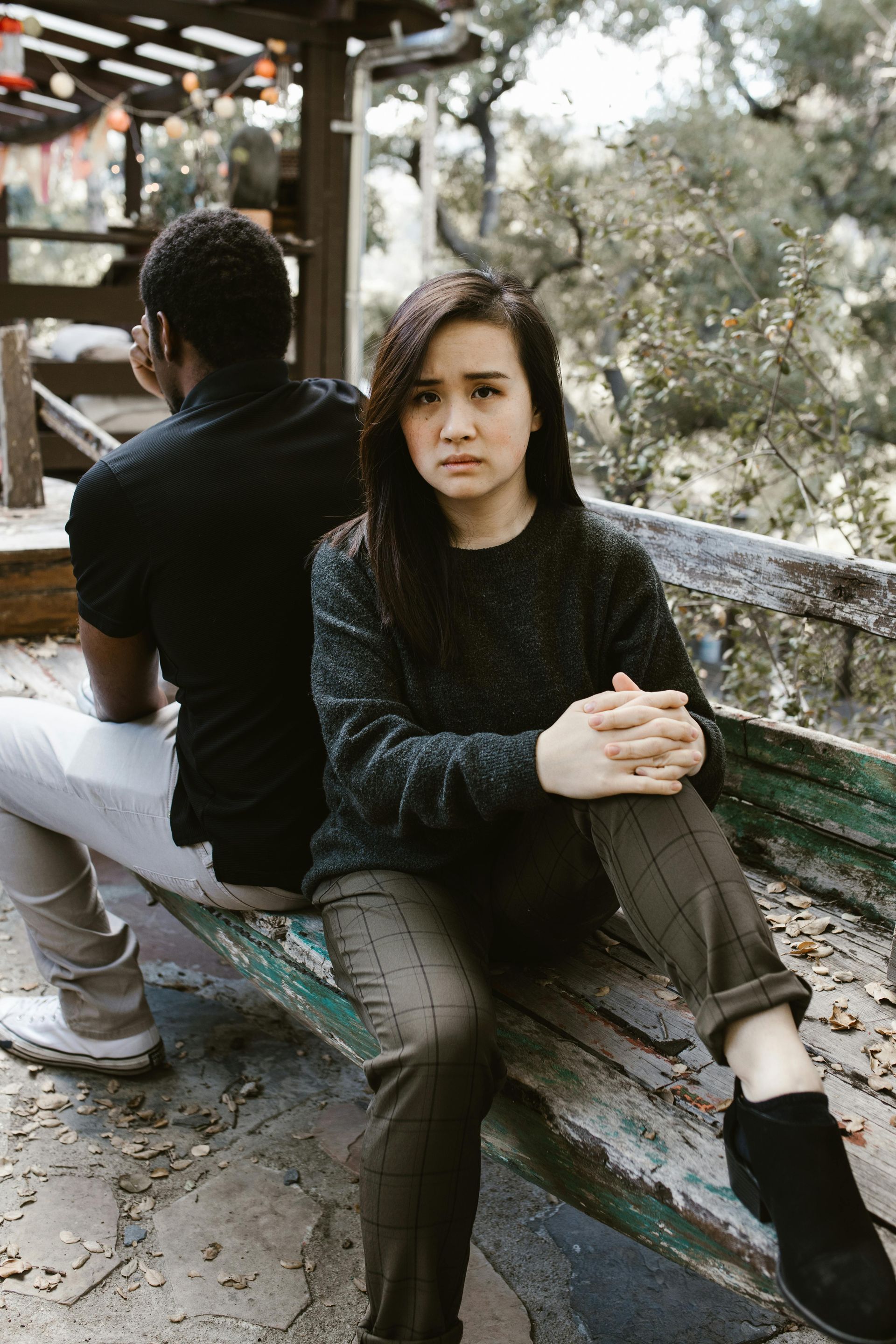 Woman with worried expression sits near a man with his back turned. Outdoors, wooden structure.