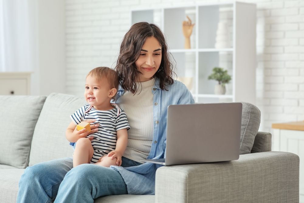 A woman is sitting on a couch holding a baby and using a laptop.
