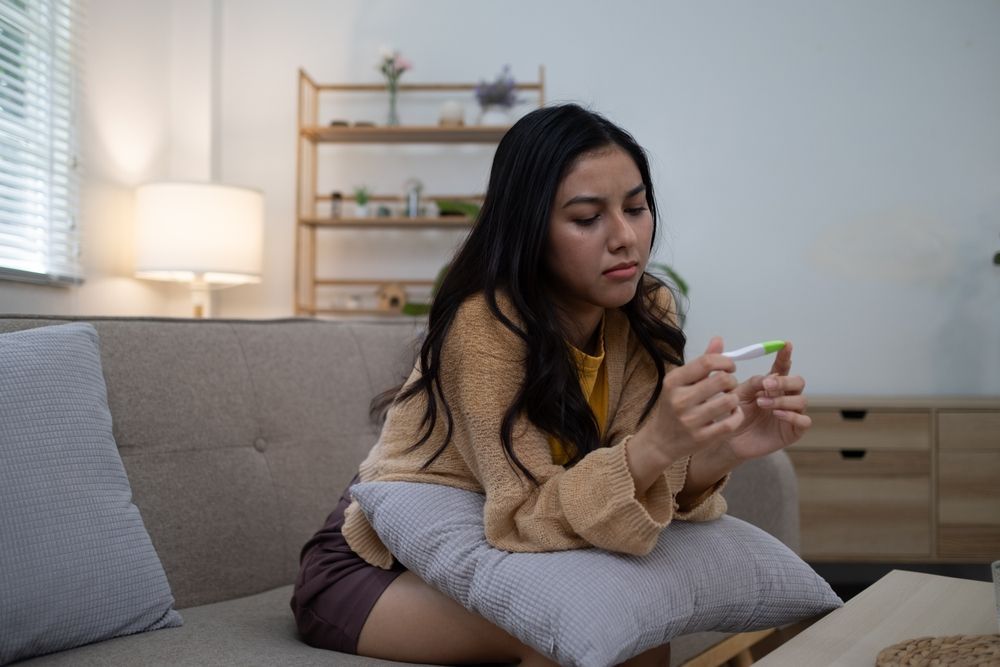 Woman looking pensive while holding a pregnancy test on a couch at home.
