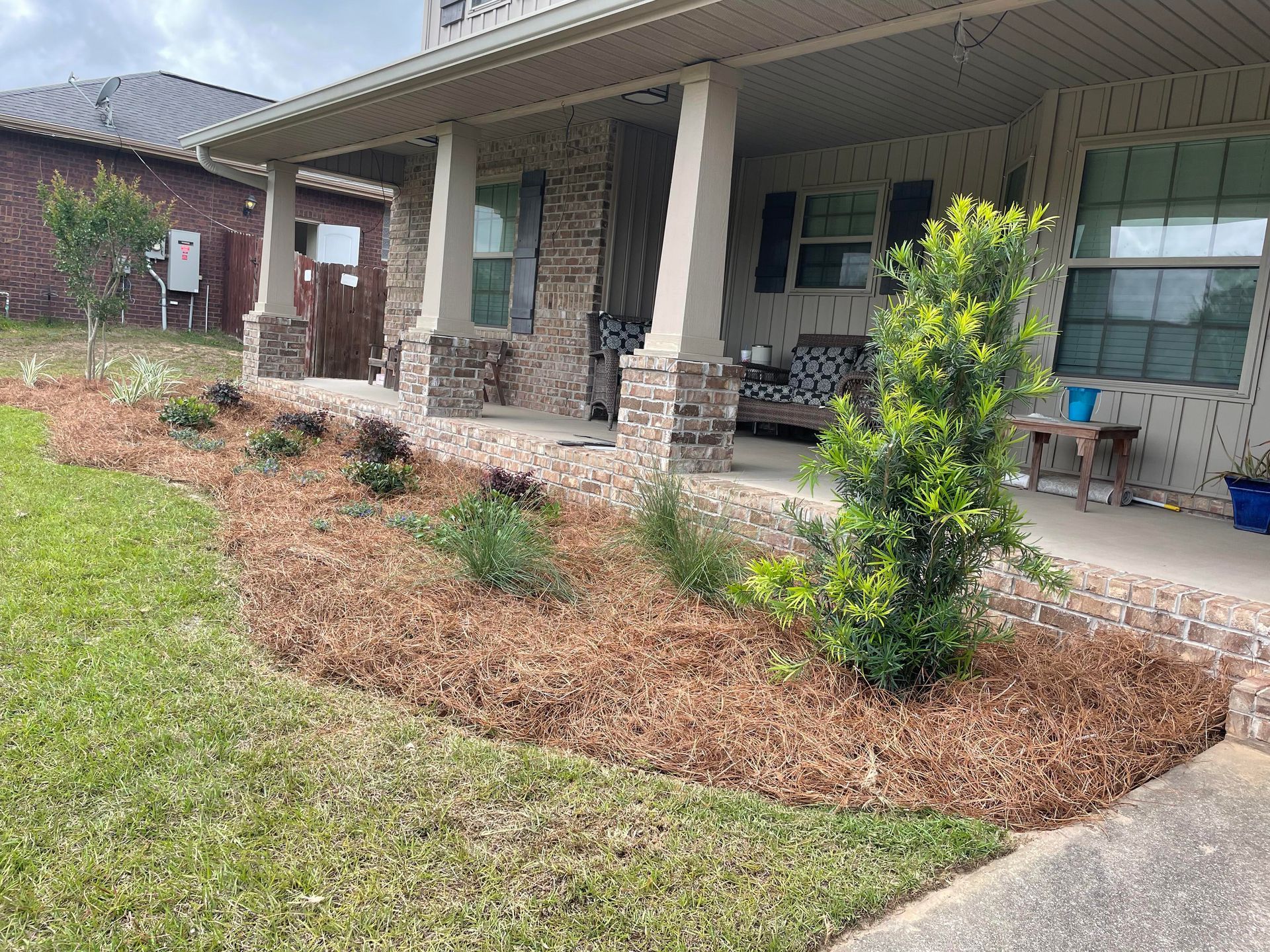 Front porch of a brick house with stone columns, a landscaped mulch bed, and a concrete walkway