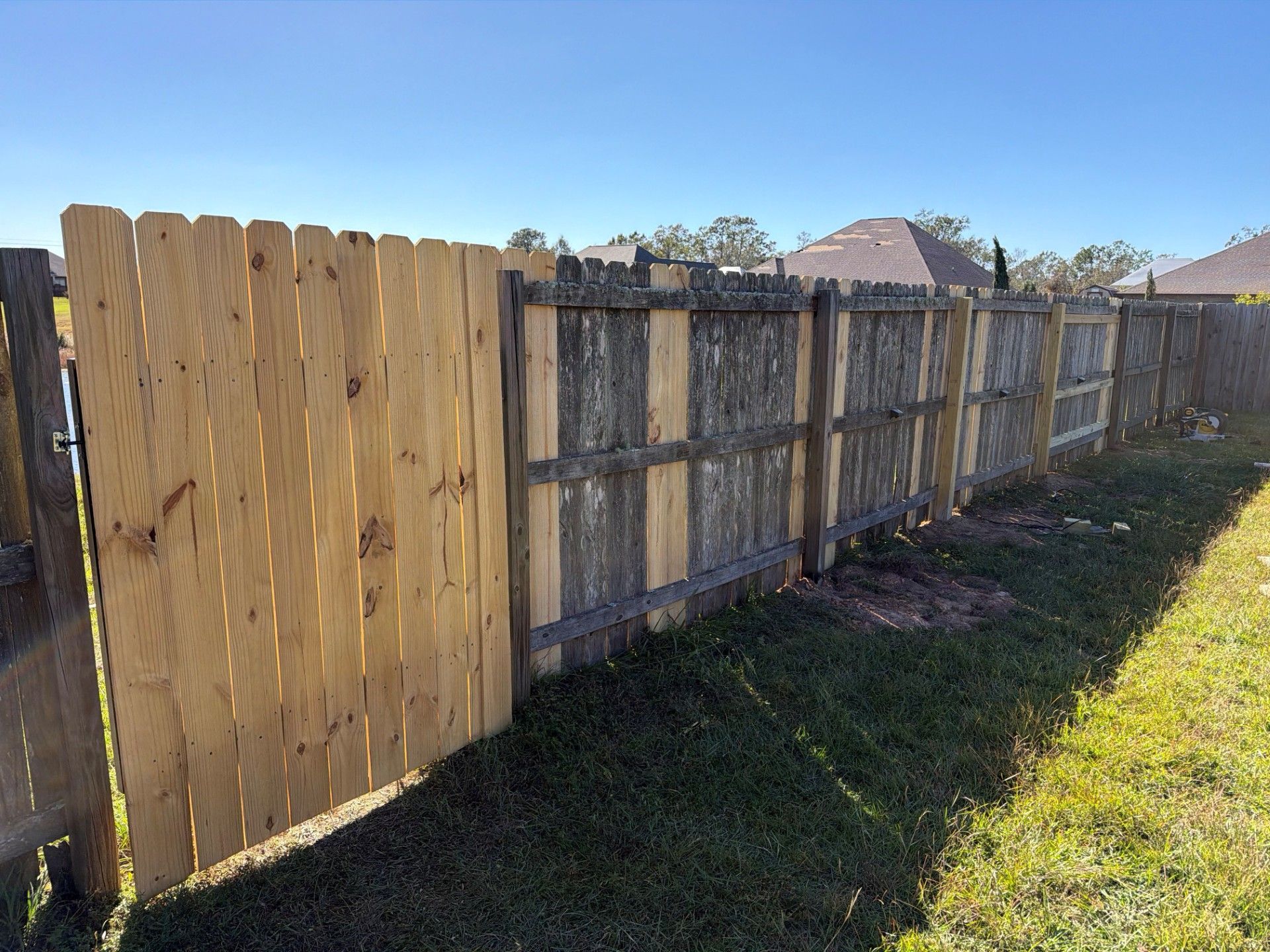 Wooden backyard fence with a new tan gate and weathered gray panels along a grassy yard.
