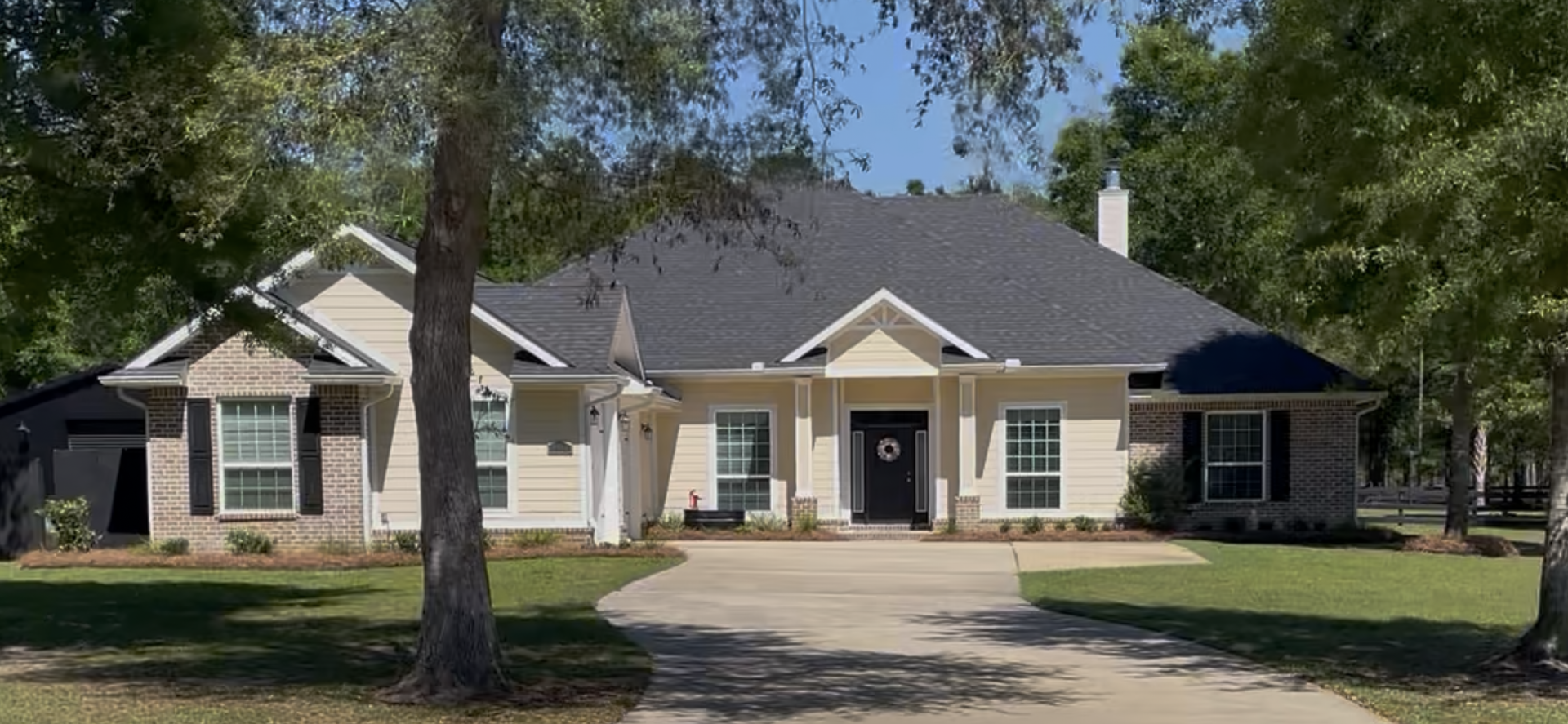 Single-story beige house with dark roof, front porch, and trees in the yard