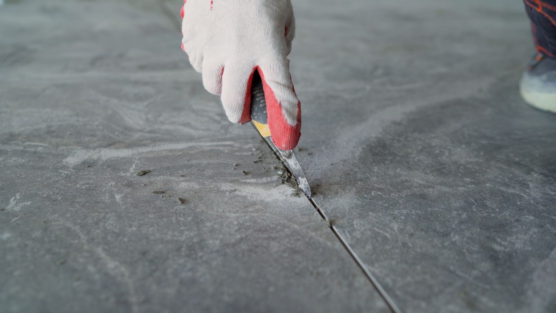 Gloved hand uses a utility knife to remove old grout from a gray tile floor.