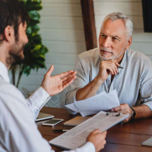 Two people discuss documents at a wooden table in a bright office meeting room.