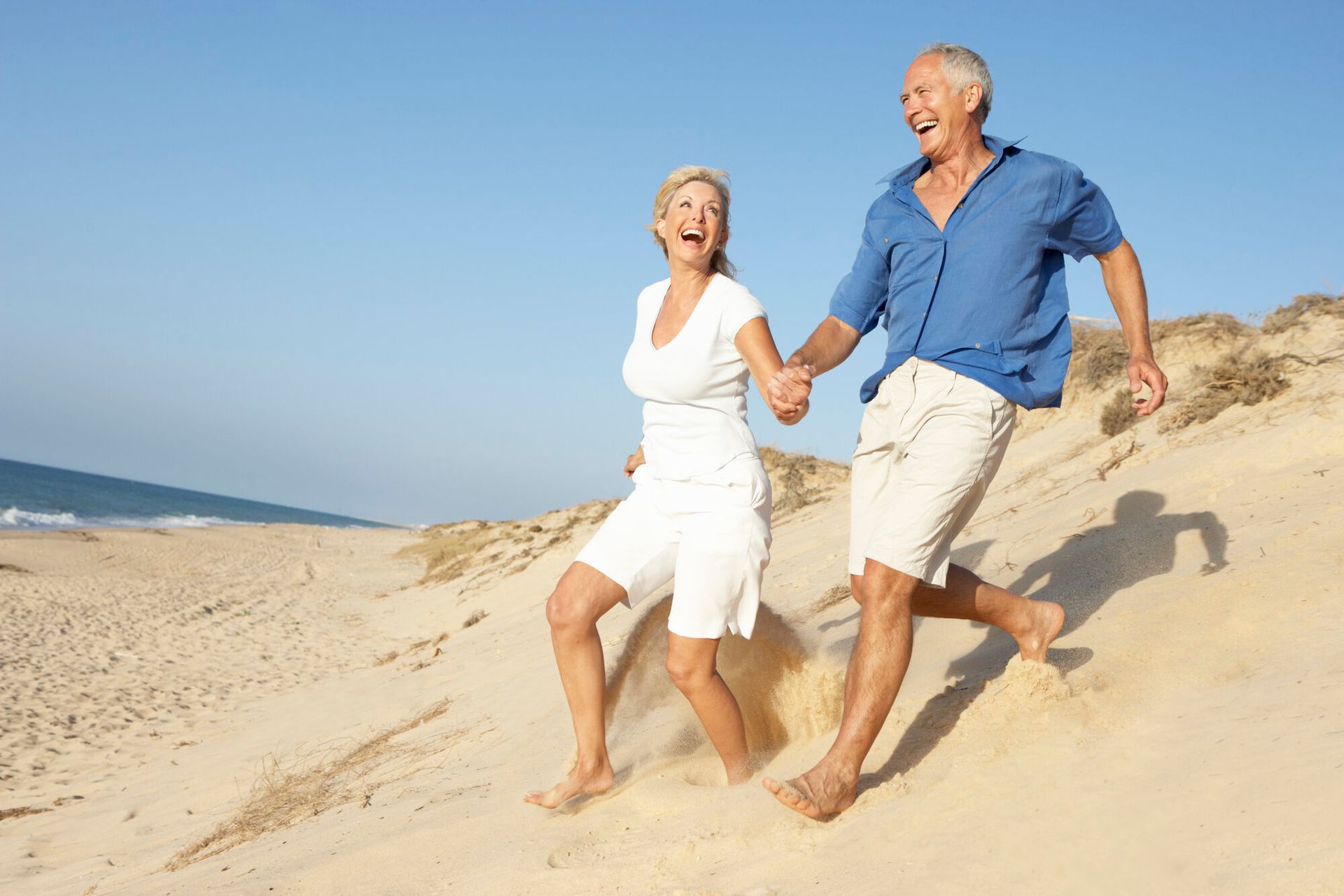Two people in white and blue clothes holding hands, smiling on a sunny beach dune