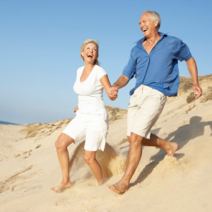 Two people in light clothing laughing and holding hands while running barefoot on sandy dunes under a blue sky