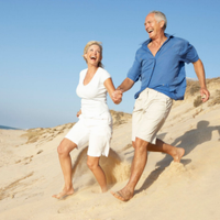 Two people in light clothing laughing and holding hands while running barefoot on sandy dunes under a blue sky