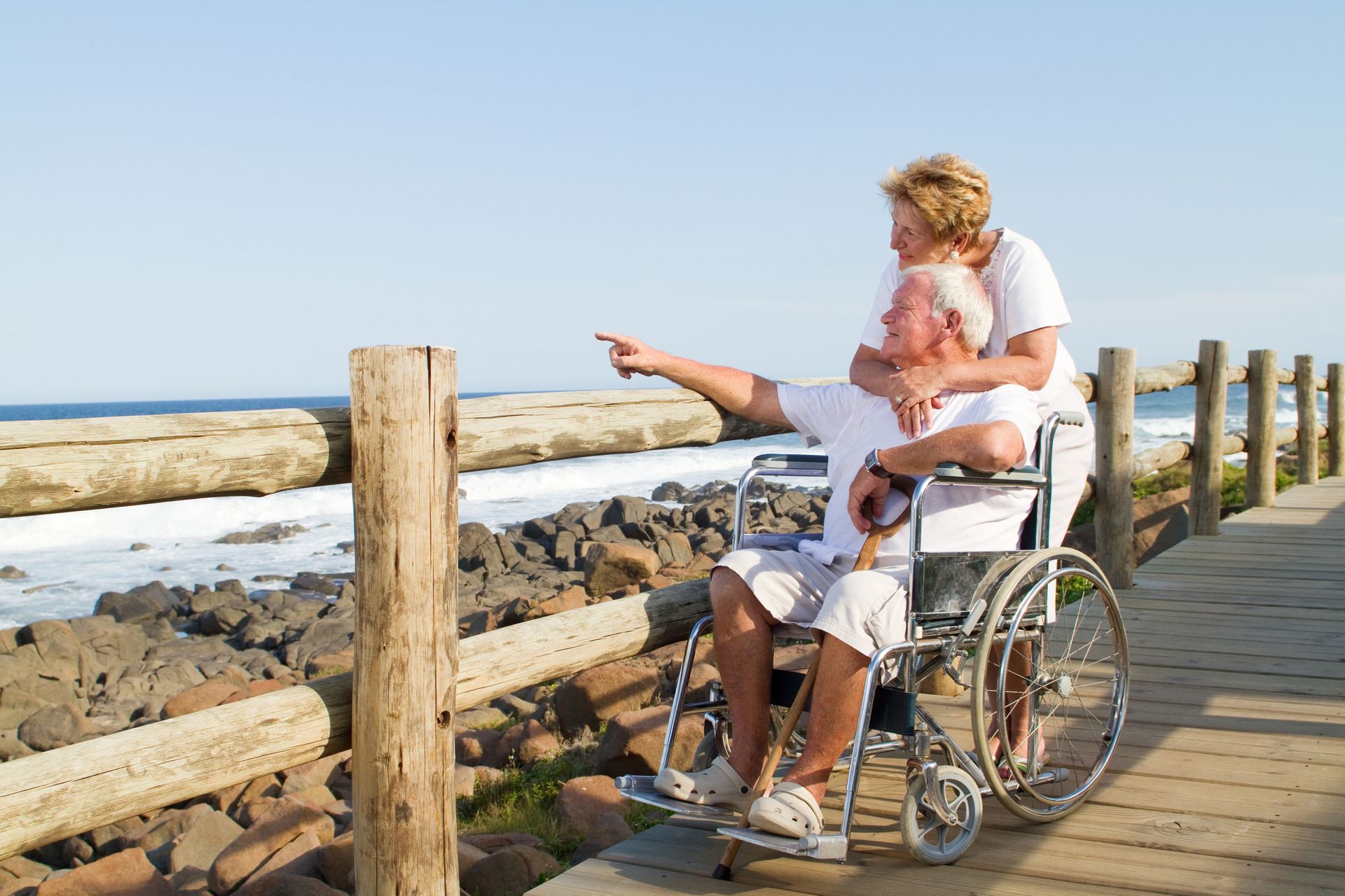 Person in wheelchair being pushed along a boardwalk by the ocean, pointing toward the shore.