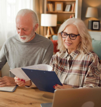 Two people reviewing papers at a wooden table in a cozy home office, smiling beside a laptop.