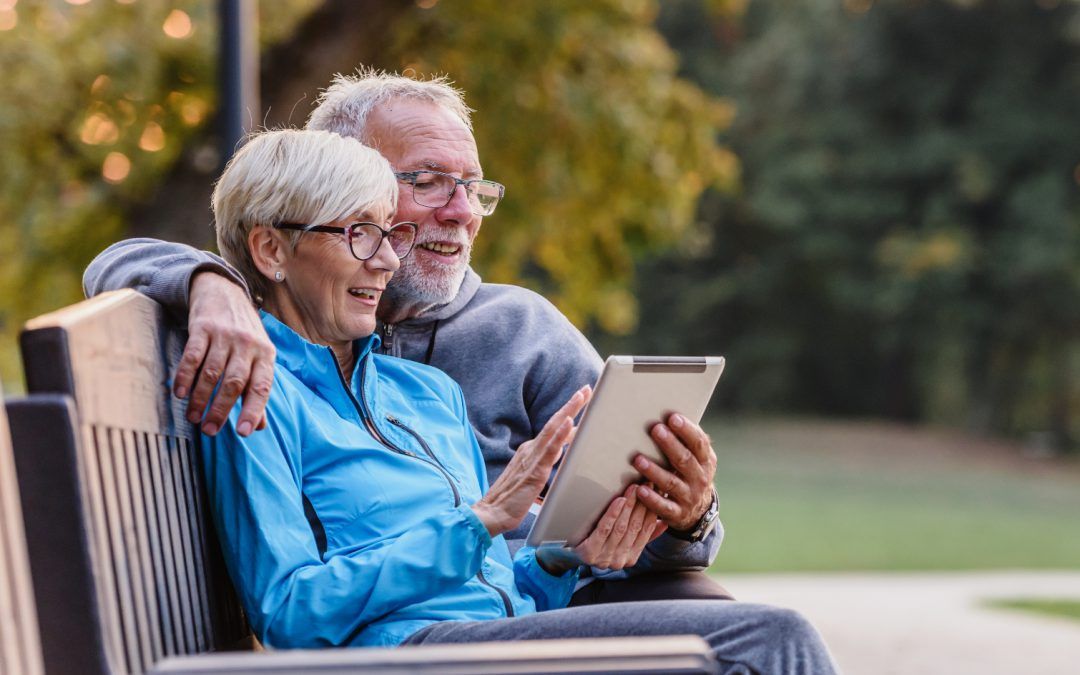 Two older adults sit on a park bench, smiling and looking at a tablet together.