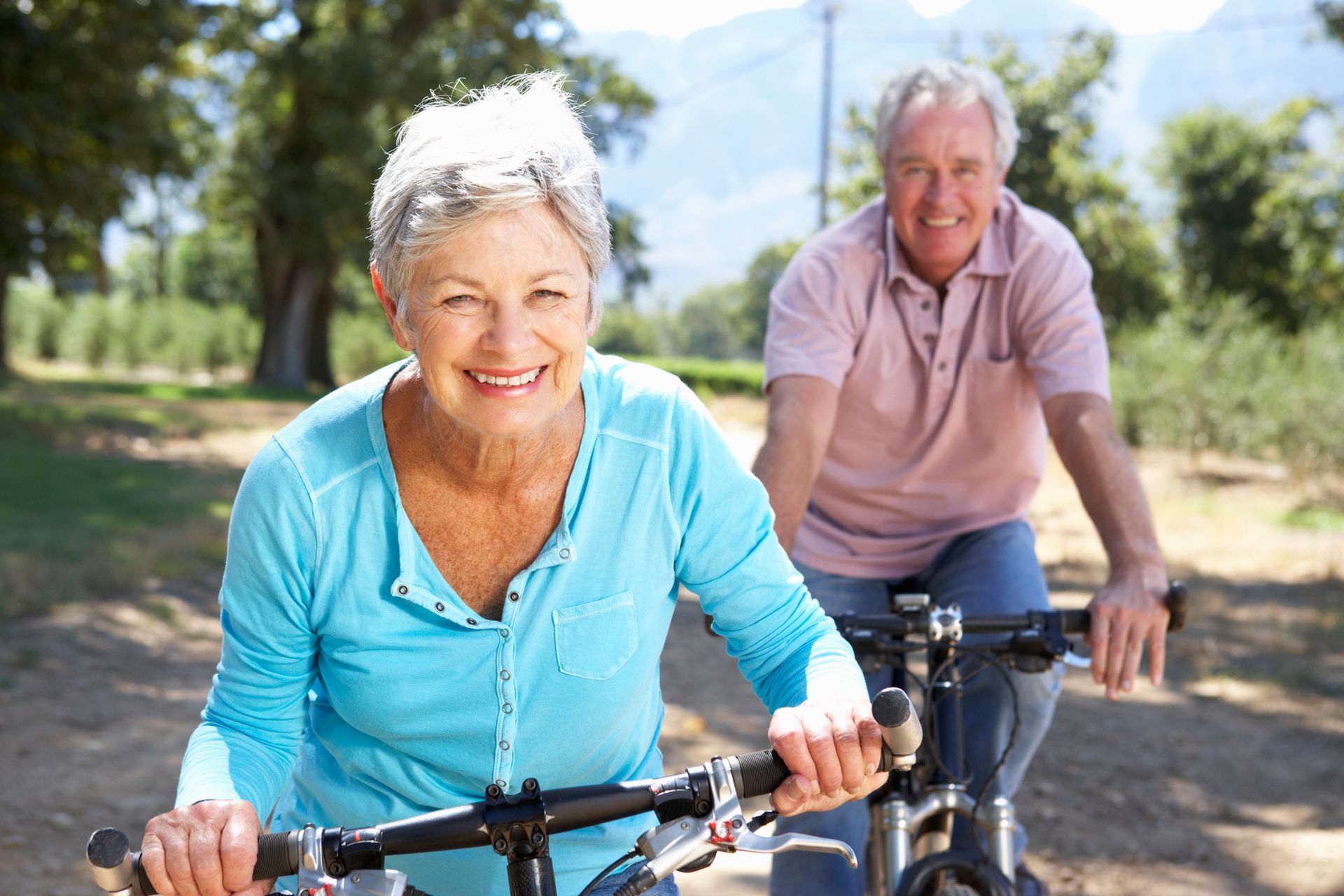 Two cyclists smiling on a sunny dirt path, riding through a green park