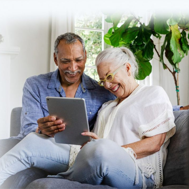 Two older adults smiling and using a tablet on a couch in a bright living room