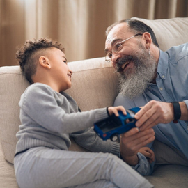 Smiling older man and child sitting on a couch, sharing a game controller in a cozy living room.