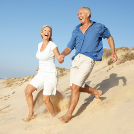 Two people in summer clothes running barefoot on a sunny sandy beach, smiling and holding hands.