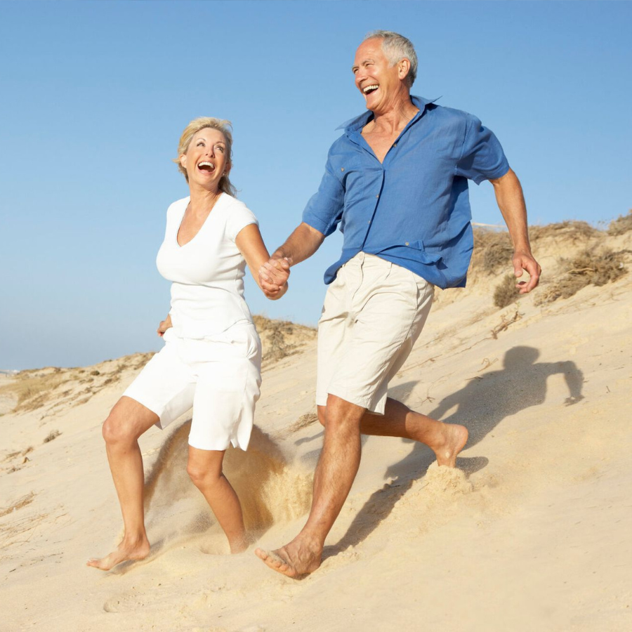 Two smiling adults holding hands, running barefoot down a sandy beach in bright sunlight.