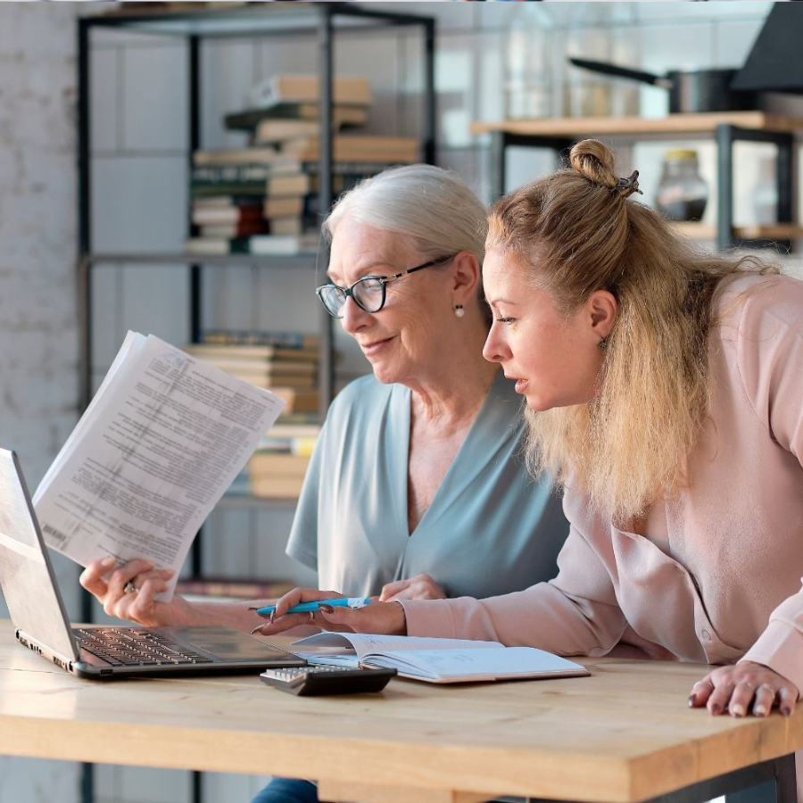 Two people reviewing documents beside a laptop at a desk in a bright office