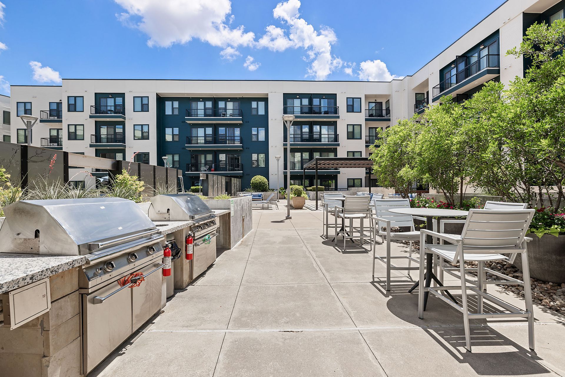 Apartment courtyard with grill stations, patio tables, and modern buildings under a bright blue sky.