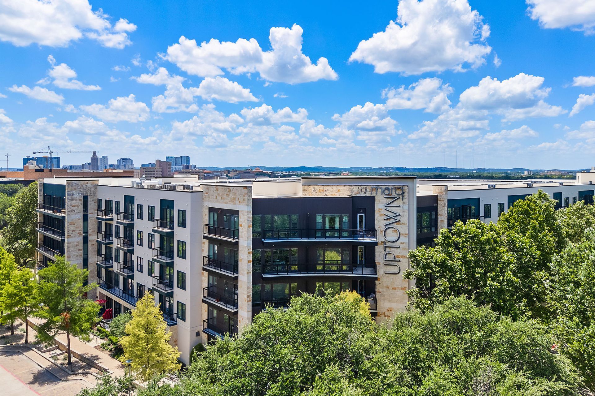Modern apartment buildings with trees under a bright blue, cloud-filled sky.