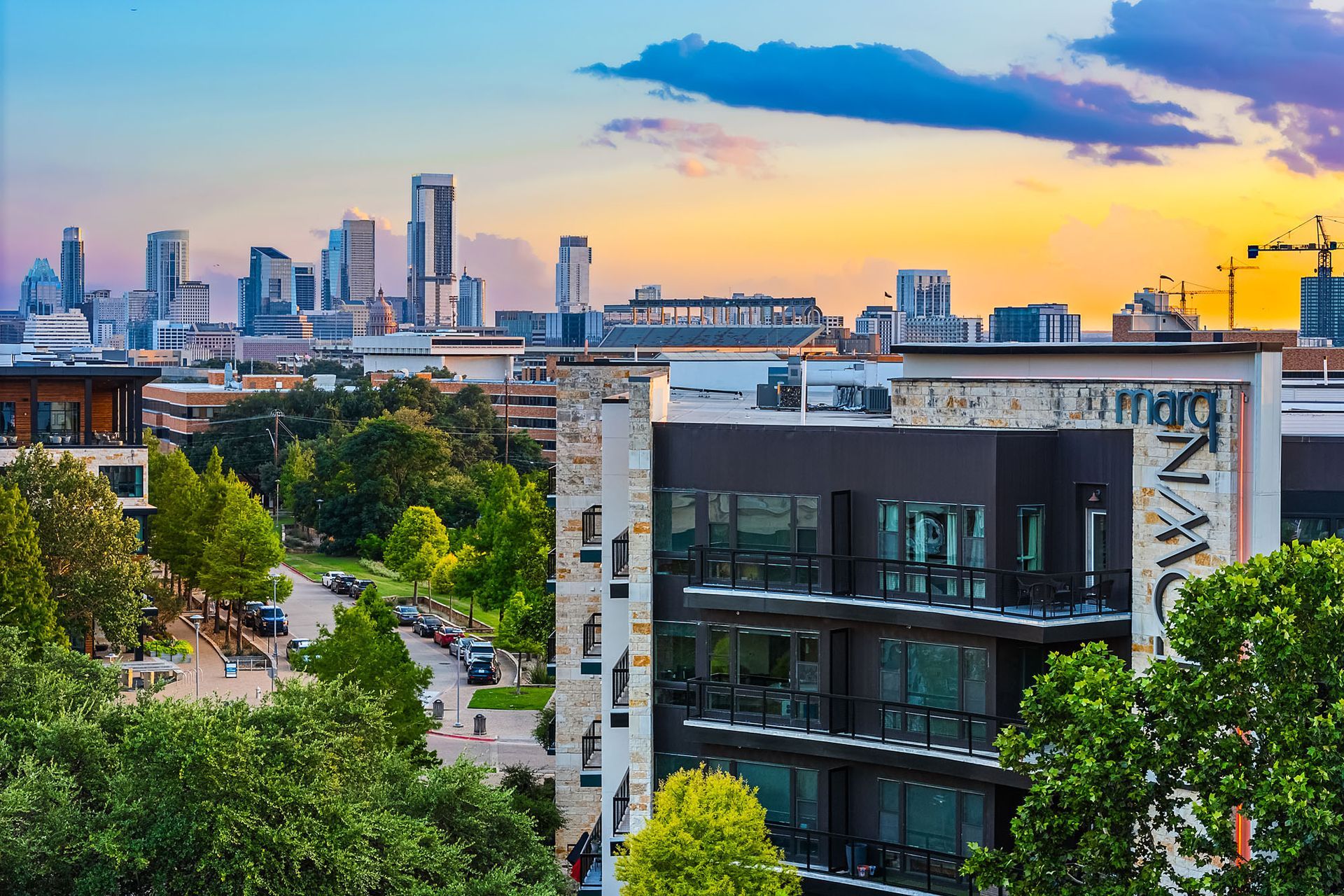City skyline at sunset beyond a tree-lined street and modern apartment building.