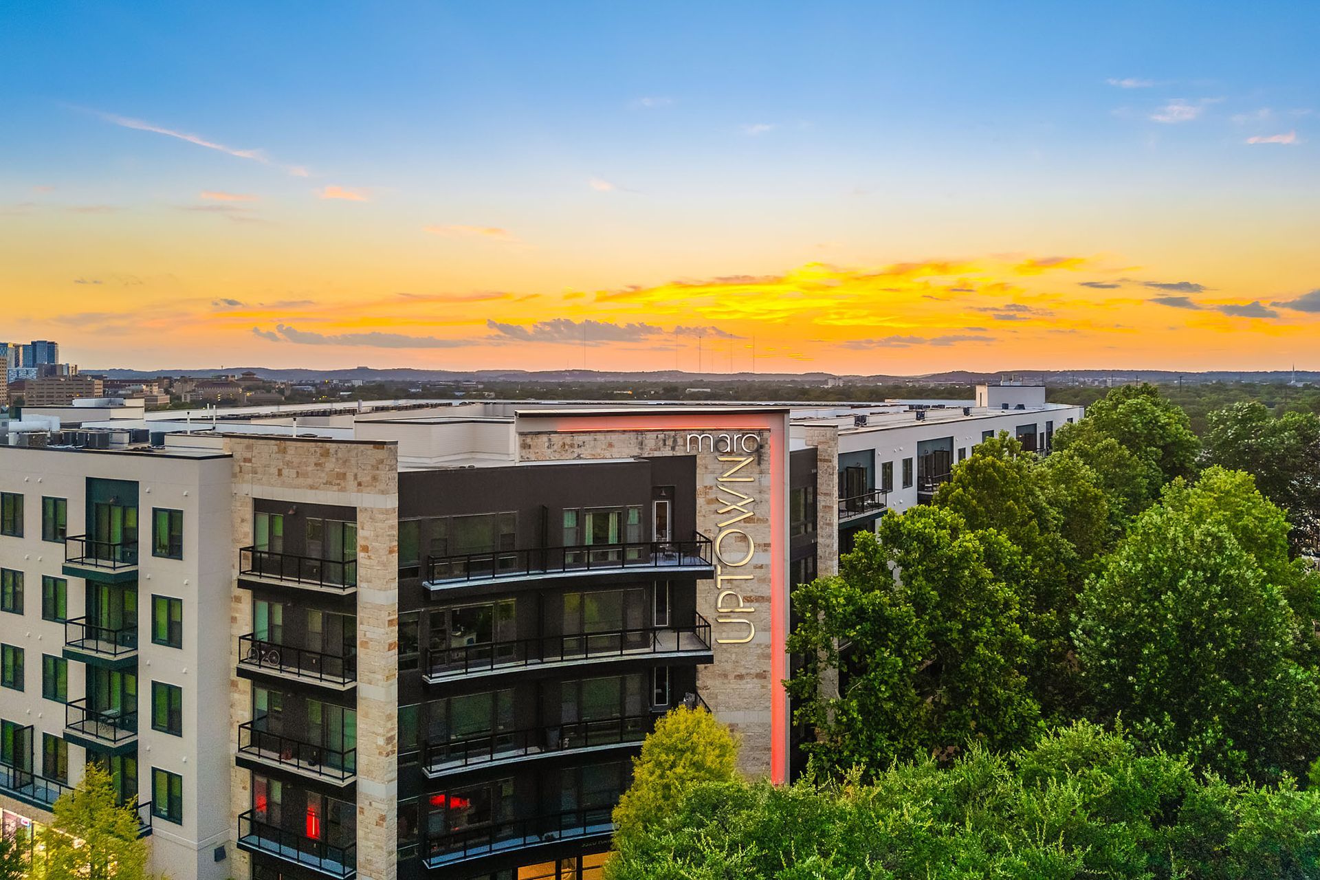 City skyline at sunset with modern buildings, green trees, and a glowing orange horizon.