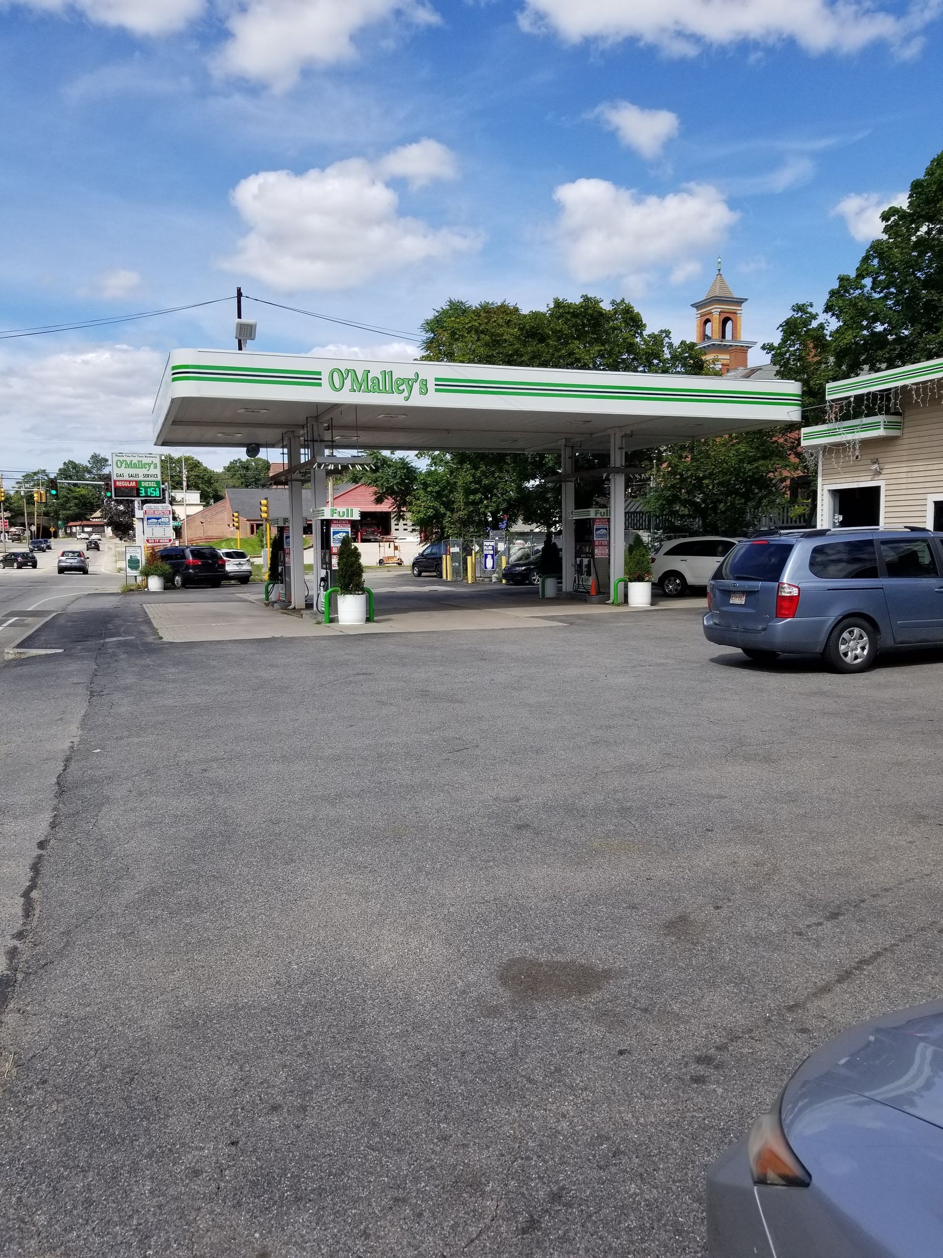 Gas station with a white and green canopy on a sunny day. A blue minivan is parked on the right.