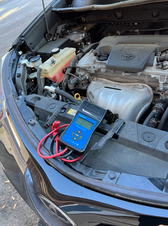 Car engine bay with a blue battery tester; red and black wires attached.