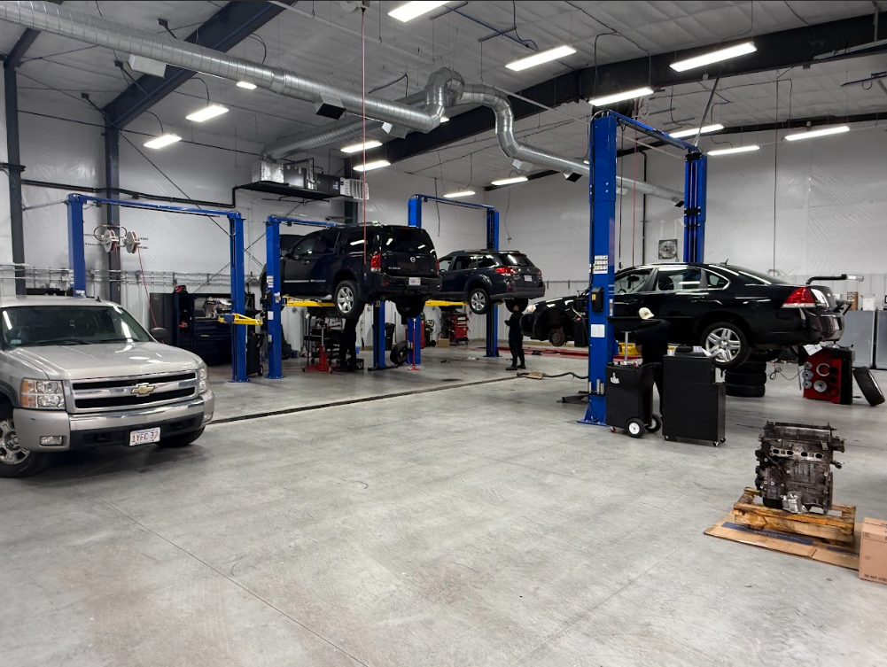 Cars on lifts inside a brightly lit auto repair shop; silver truck in foreground.