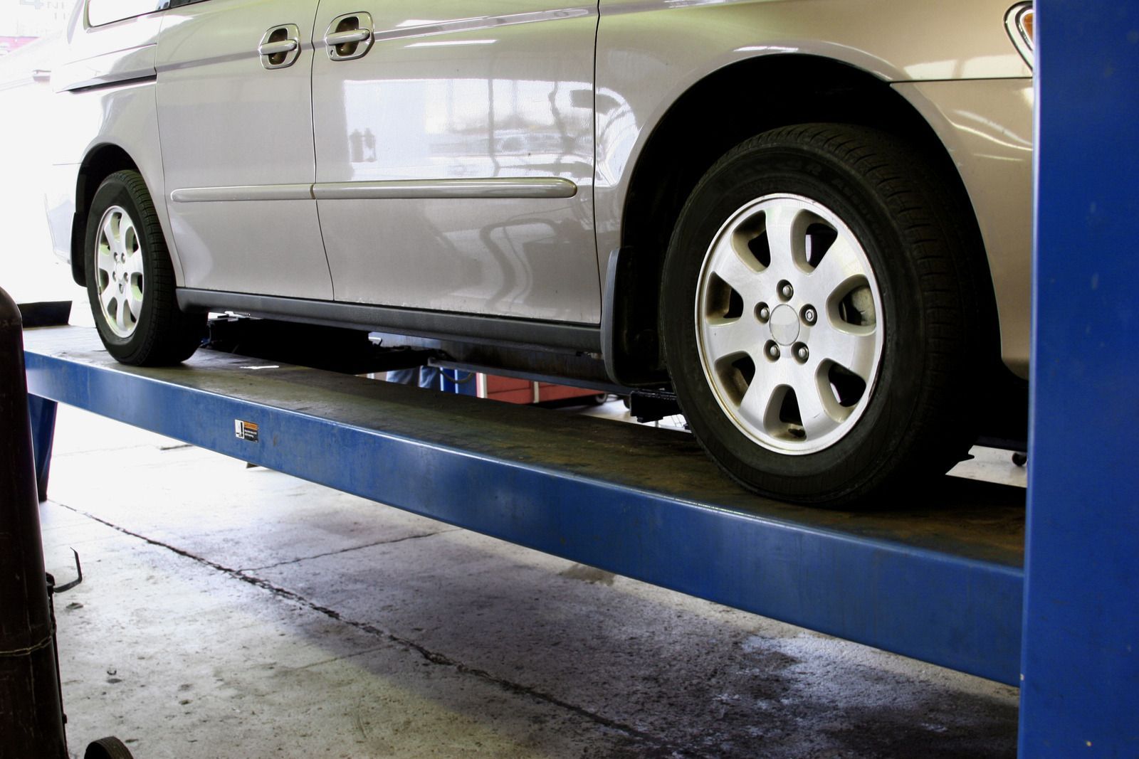 Silver minivan on a blue car lift inside a garage, tires visible, sunlight.