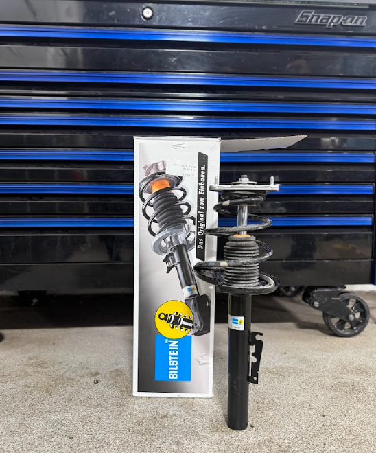 A car suspension strut and spring assembly sits next to its Bilstein box, in front of a blue and black toolbox.