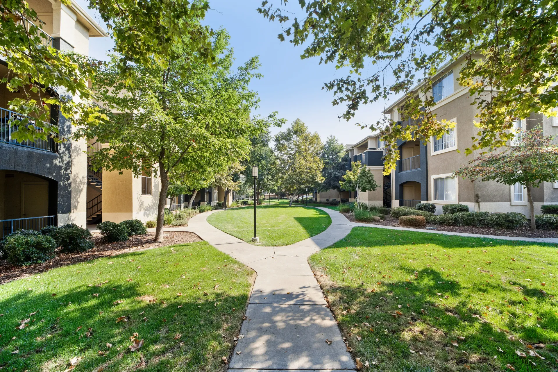 Outdoor community courtyard with trees, green lawn, and curved walkways between apartment buildings at Cross Pointe apartments in Antioch, CA.