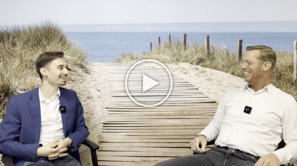 Twee mensen zitten op een strandpromenade, lachend en pratend, met een afspeelicoontje eroverheen.