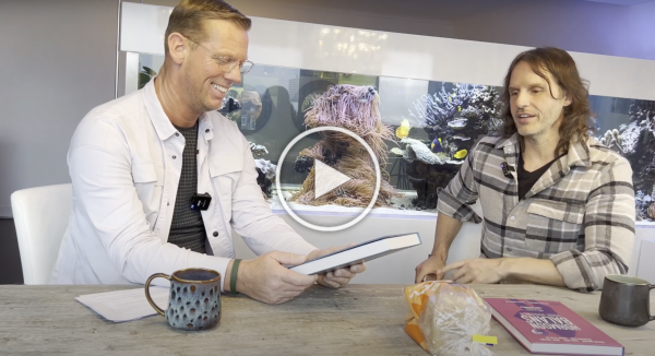 Twee mensen praten aan een tafel met een mok en boeken, met een video-afspeelknop boven de scène.