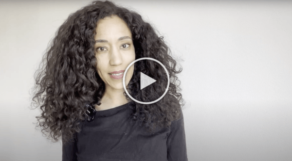 Vrouw met krullend haar lacht tegen een grijze achtergrond, met een afspeelknop eroverheen.