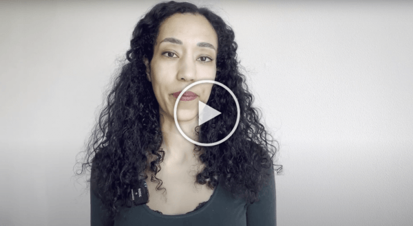 Vrouw met krullend zwart haar spreekt tegen een lichtgrijze achtergrond, met een afspeelknop eroverheen.