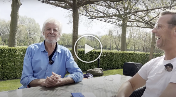 Twee mensen die met elkaar praten aan een picknicktafel onder een afdak in een tuin, met een afspeelknop op de achtergrond.