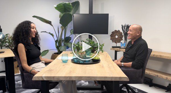 Twee mensen zitten tegenover elkaar aan een tafel in een moderne vergaderruimte, met een plant en een monitor op de achtergrond.