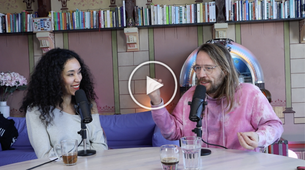 Twee mensen zitten in een podcaststudio en spreken in microfoons aan een tafel met boekenplanken achter hen.