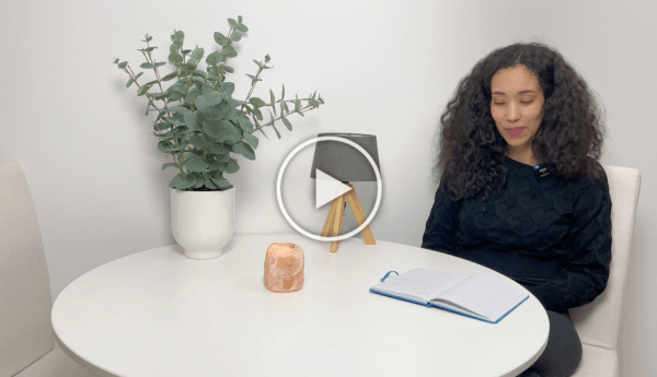 Vrouw zit aan een witte tafel met een notitieboekje, een plant, een kaars en een pictogram van een spelletje op de tafel.