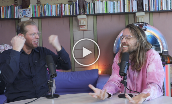 Twee mensen in een podcaststudio, de een spreekt in een microfoon terwijl de ander gebaart, met boekenplanken achter hen.