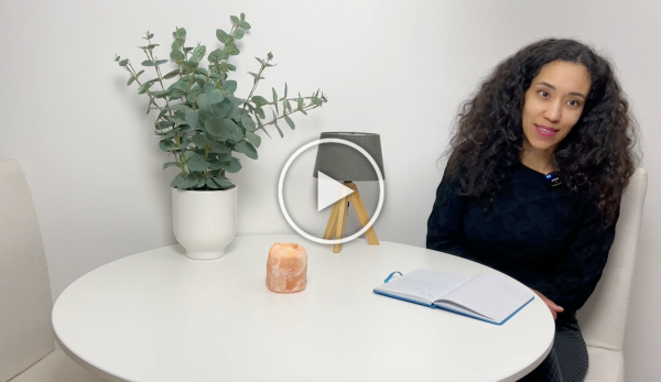 Vrouw zit aan een witte ronde tafel met een plant, een kaars en een open notitieboekje in een lichte kamer.