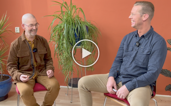 Twee mensen zitten te praten in een kamer met een oranje muur, een potplant en een overlay van een video-afspeelknop.
