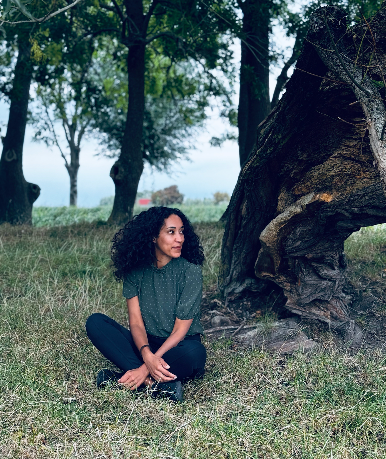 Vrouw met krullend haar zit met gekruiste benen onder een grote boom in een grasveld.