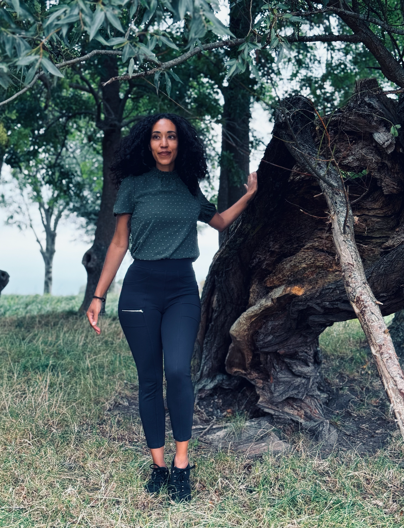 Vrouw staat bij een grote boom in een grasveld, gekleed in een donkere top en legging.
