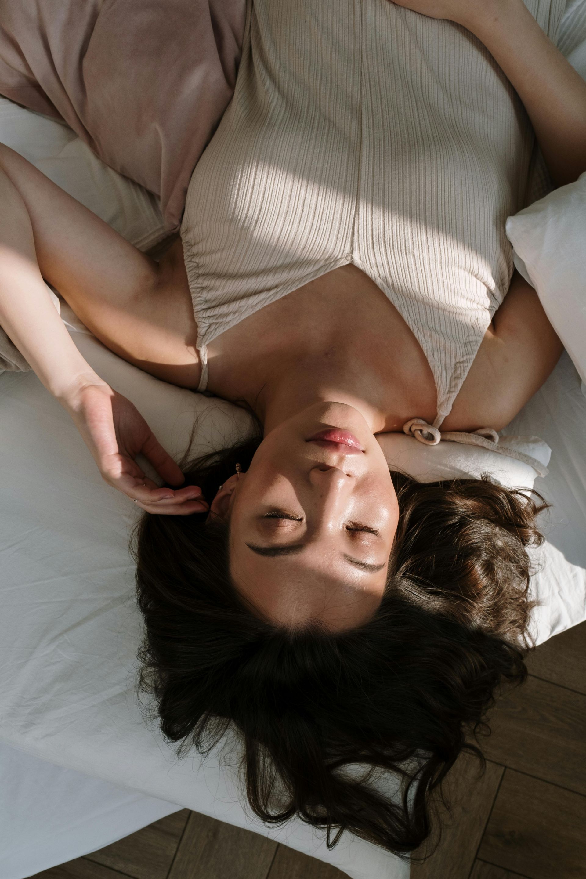 Vrouw ligt op bed in een witte gebreide top en roze broek, ogen gesloten in zacht natuurlijk licht.