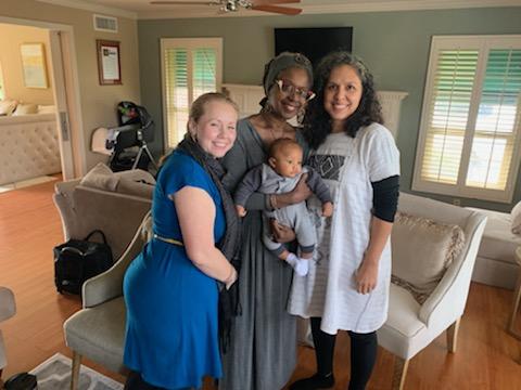 Four people smiling, posing with a baby in a living room. The woman in the center is holding the baby.
