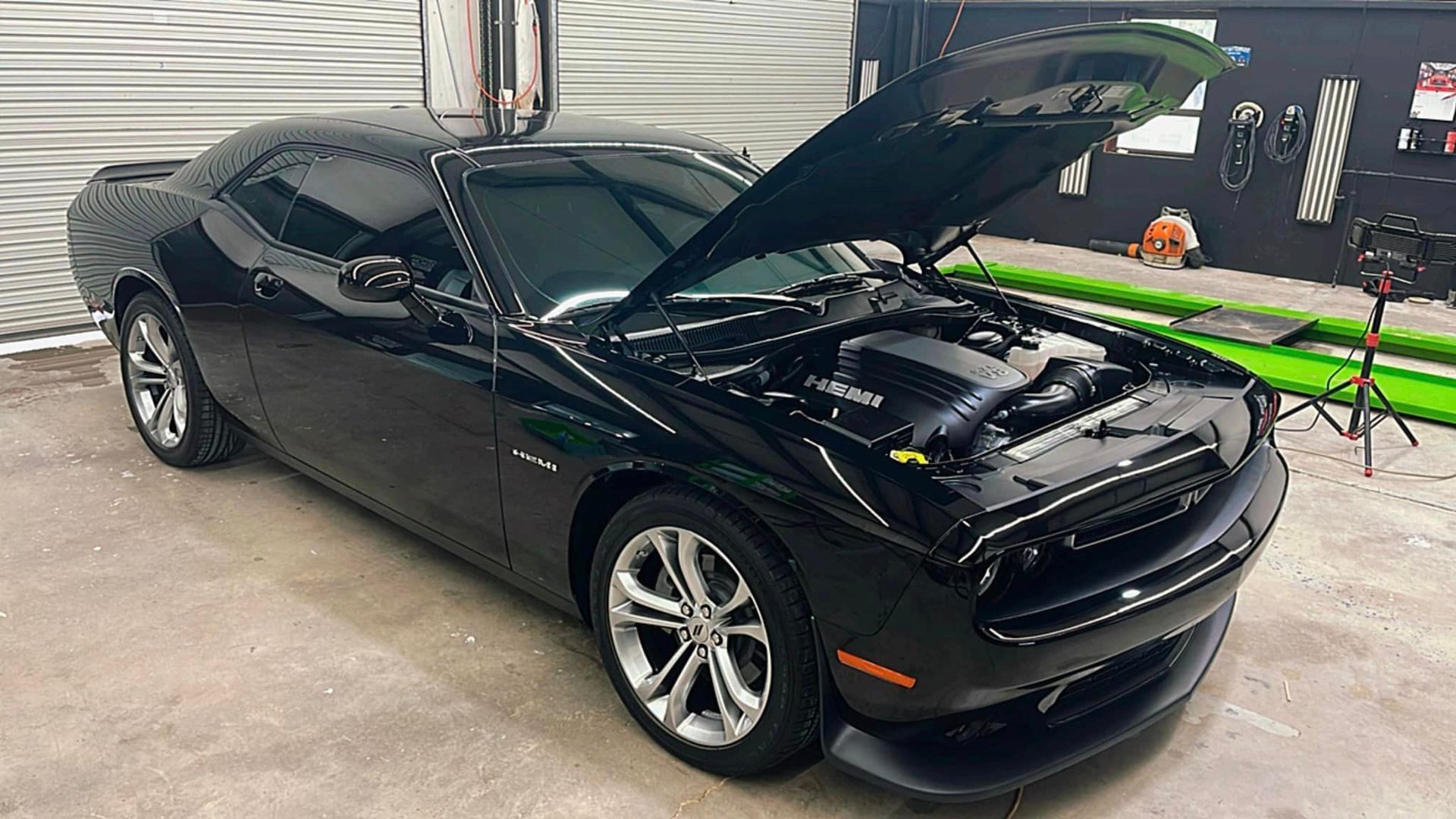 A black dodge challenger with the hood up is parked in a garage.